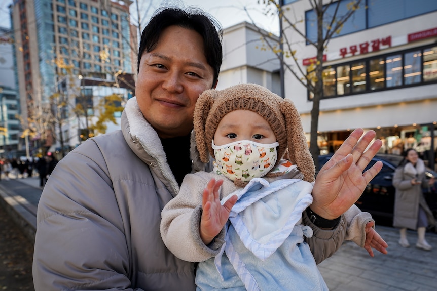 A man smiles and waves while holding a child wearing a mask and hat with long fuzzy ears