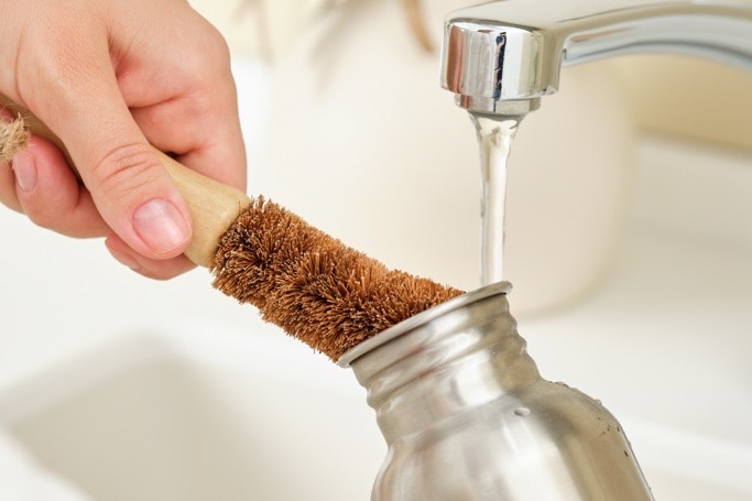 Uma garrafa de água de metal é lavada em água corrente com uma escova marrom.