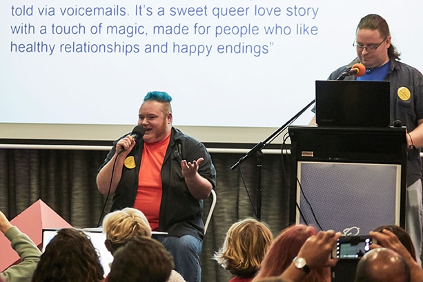 A man w/blue hair sitting on a chair and a man standing up both speaking into microphones in front of a powerpoint presentation