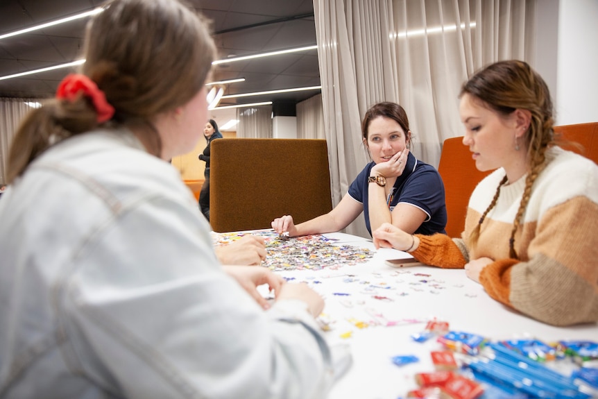 A woman talks to two female students over a half completed puzzle.
