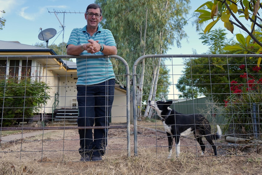 A man stands outside his home, leaning on a gate next to a dog. 