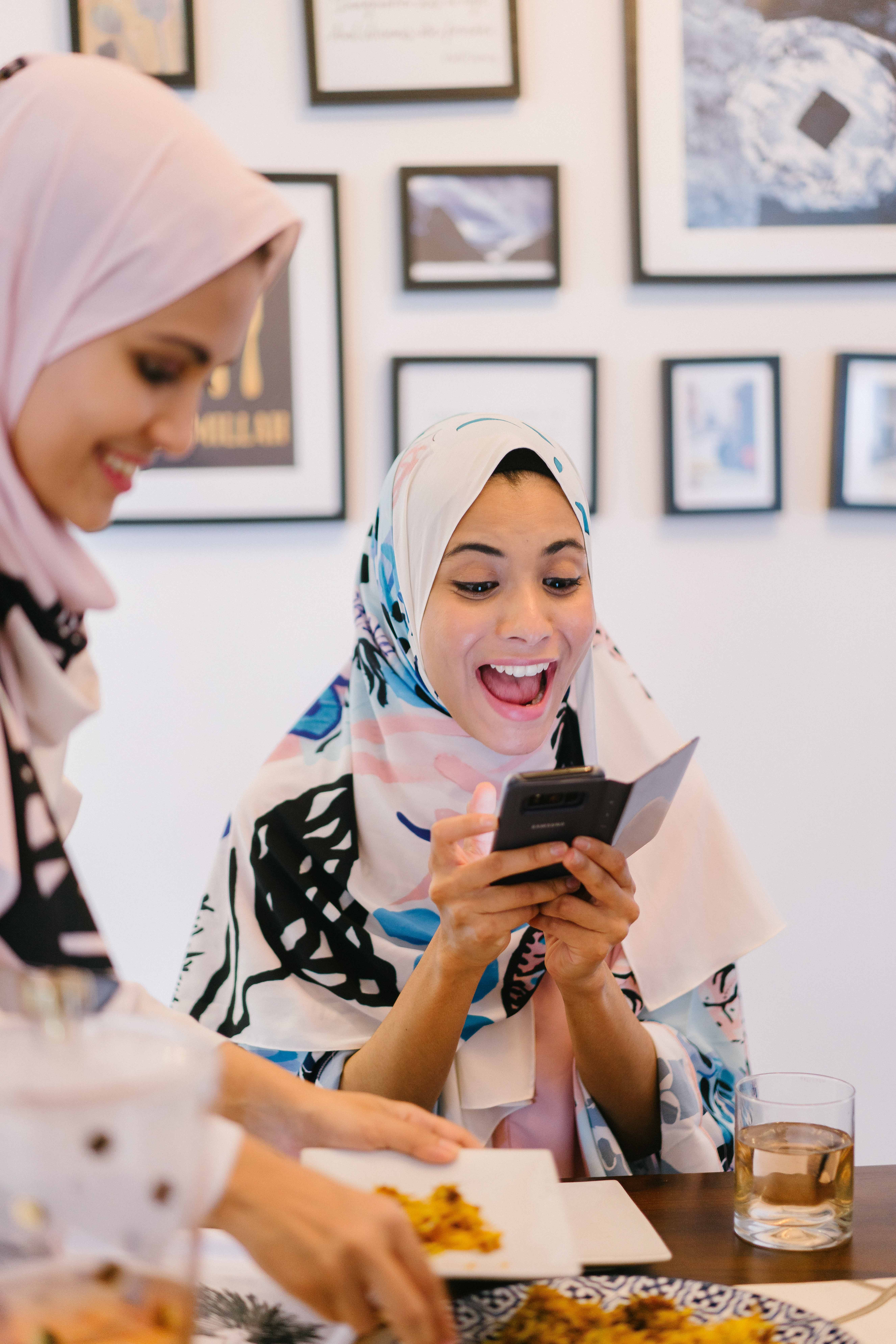 Two women wear hijabs while one women laughs and looks at her mobile phone to depict making your Instagram body positive.