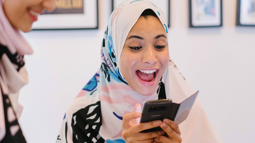 Two women wear hijabs while one women laughs and looks at her mobile phone to depict making your Instagram body positive.