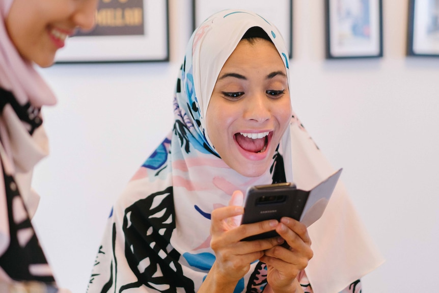 Two women wear hijabs while one women laughs and looks at her mobile phone to depict making your Instagram body positive.