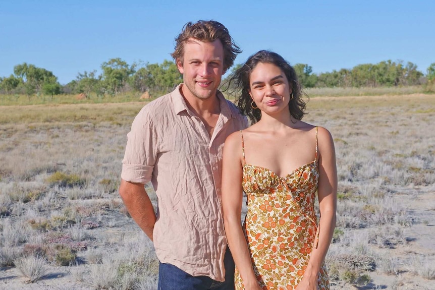 A man is seen standing behind a woman in a wide open plain, with several trees and shrubs far behind them. They both smile.