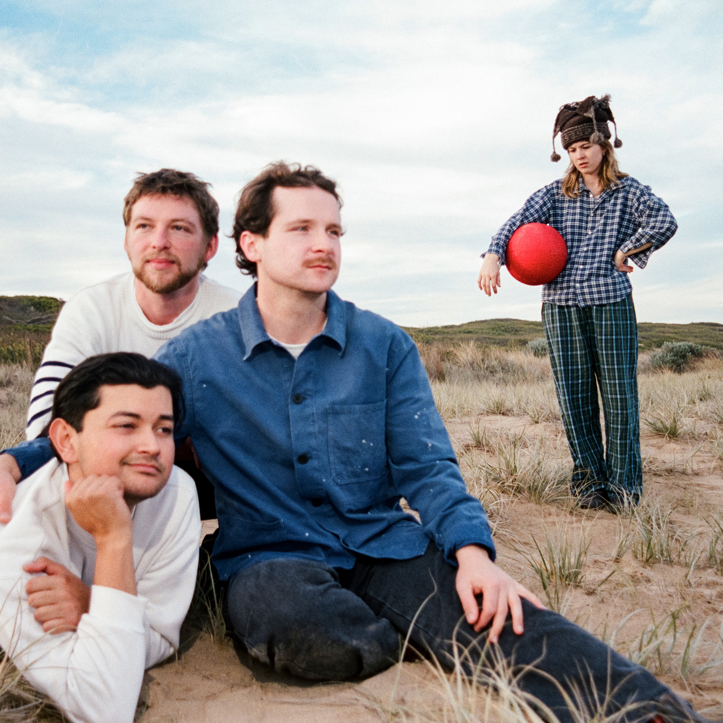 Three males are on a rock at the beach, the fourth individual is a female wearing a jester hat and holding a red ball