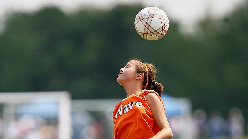 A young girl in orange heads a ball. 