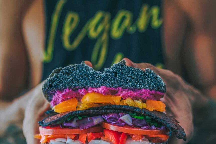 Close up of hands holding a burger filled with brightly coloured vegetables. In the background, the person's shirt reads "Vegan"