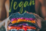 Close up of hands holding a burger filled with brightly coloured vegetables. In the background, the person's shirt reads "Vegan"
