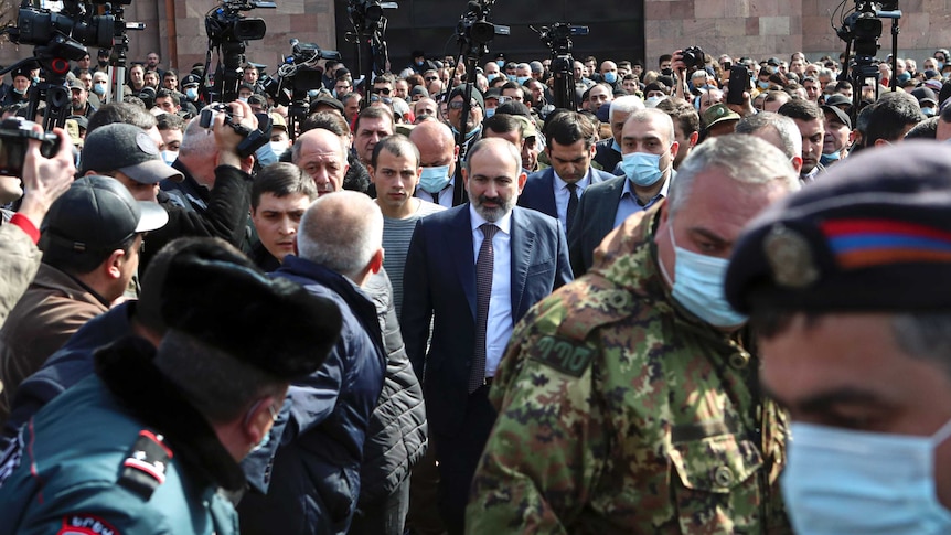 A man in a suit stands amidst a large crowd including men in military uniforms and media.
