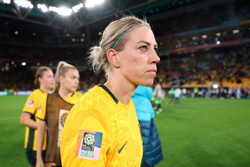 A soccer player wearing yellow and green forlornly looks at the crowd with team-mates behind her after a game