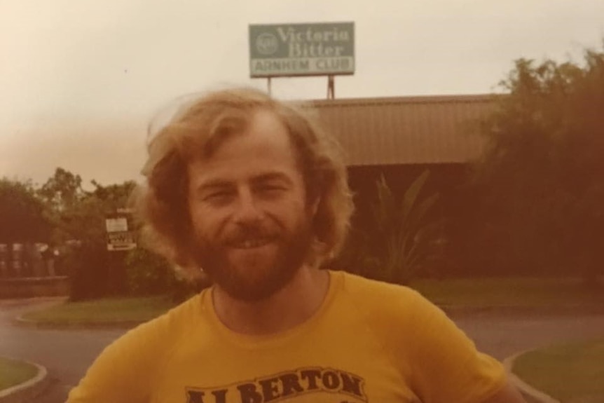 A grainy vintage photo of a young man with shaggy hair standing in front the 'Victoria Bitter Arnhem Club' sign