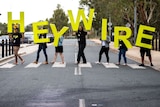 Seven people walk across a zebra crossing holding giant green letters spelling out the word HEYWIRE.