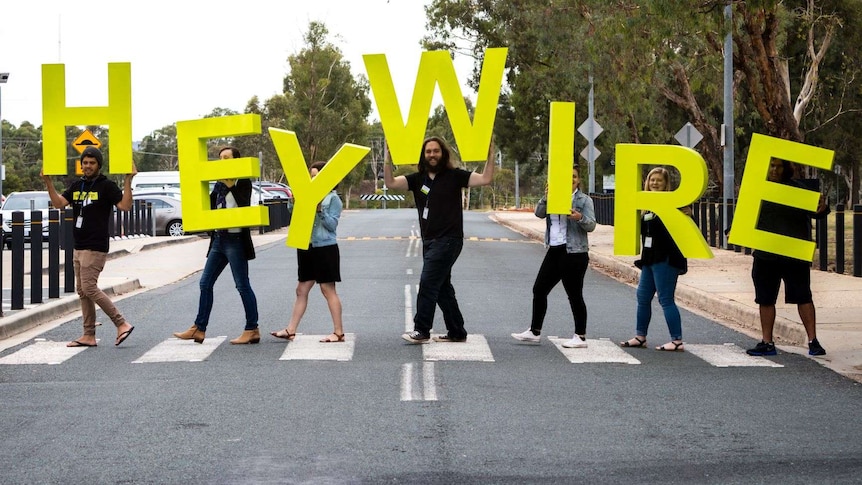 Seven people walk across a zebra crossing holding giant green letters spelling out the word HEYWIRE.