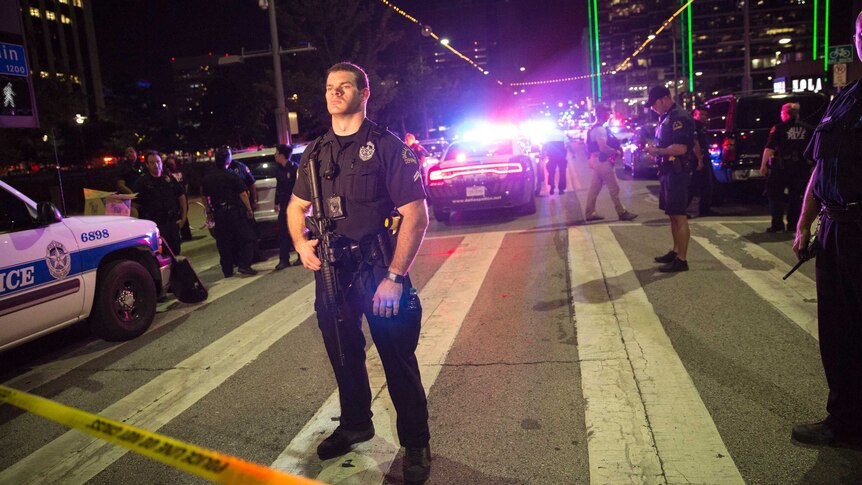 A police officer stands in the street holding a rifle. Police cars with lights on are in the background.