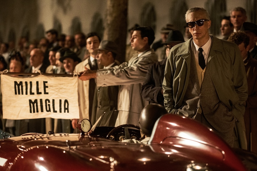 A film still of Adam Driver, with white hair and sunglasses, hands in the pockets of his trench coat, beside a vintage car.