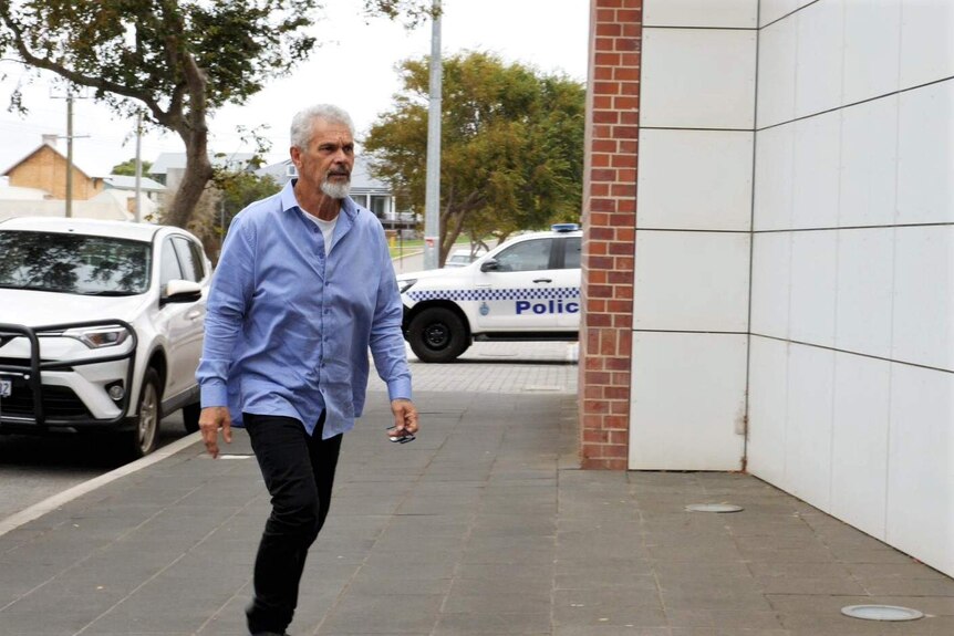 Grey-haired man in blue shirt walks towards building looking serious with police cars in the background.