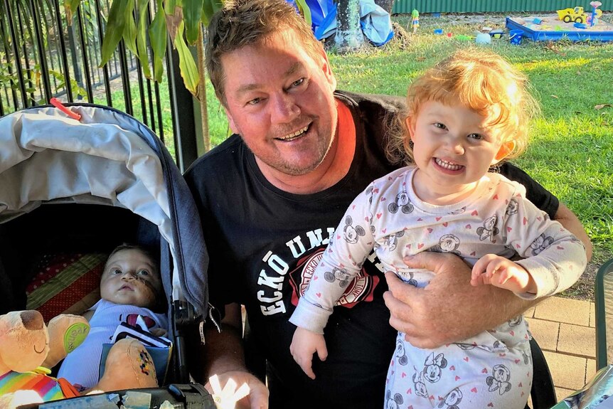A smiling dad holds his two-year-old daughter in a backyard while a baby lies in a pram.