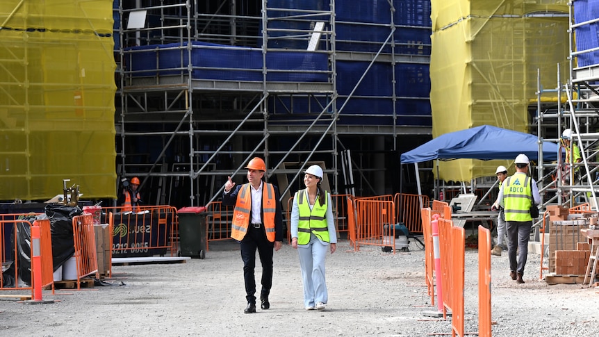 a wide shot of cameron dick and meagan scanlon wakling through a construction site wearing high-vis vests