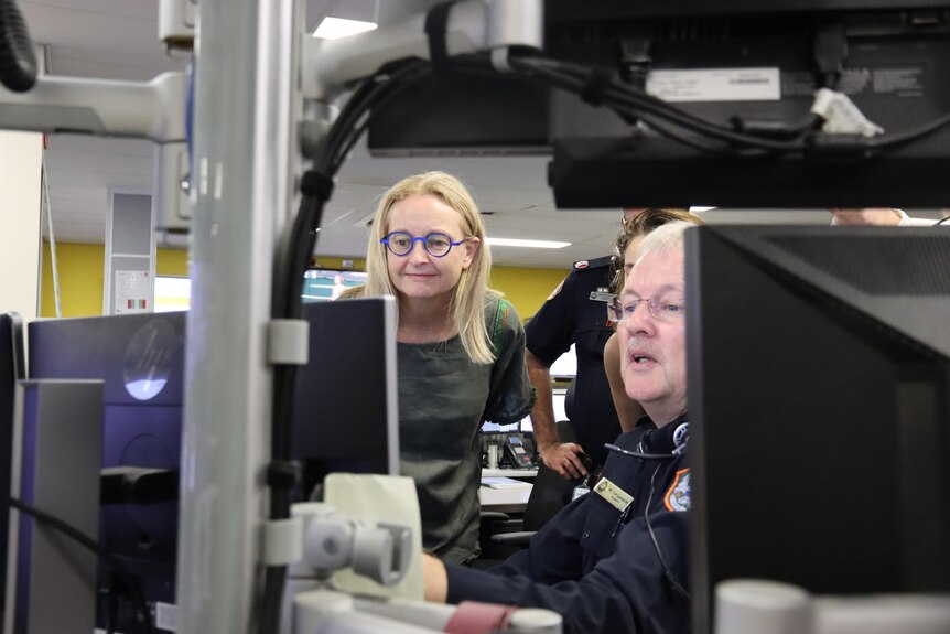 A woman with blonde hair and bright blue glasses looks over the shoulder of a police officer sitting at an office desk