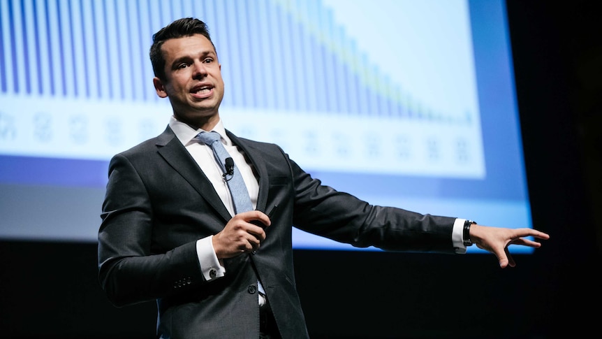 A man in a suit addresses an audience while standing in front of a graph on a power point.
