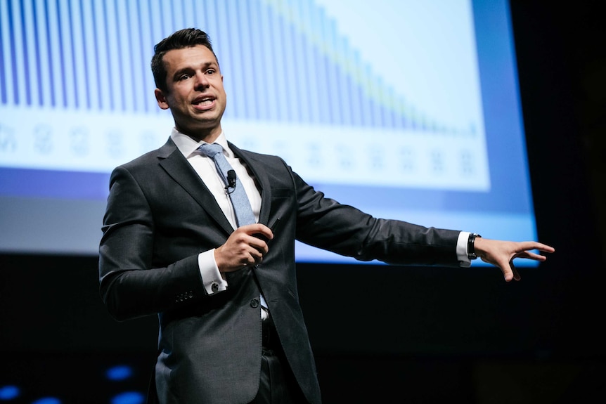 A man in a suit addresses an audience while standing in front of a graph on a power point.