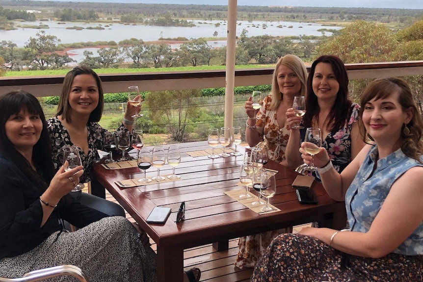 Five women sit at a table raising wine glasses with wetland in background
