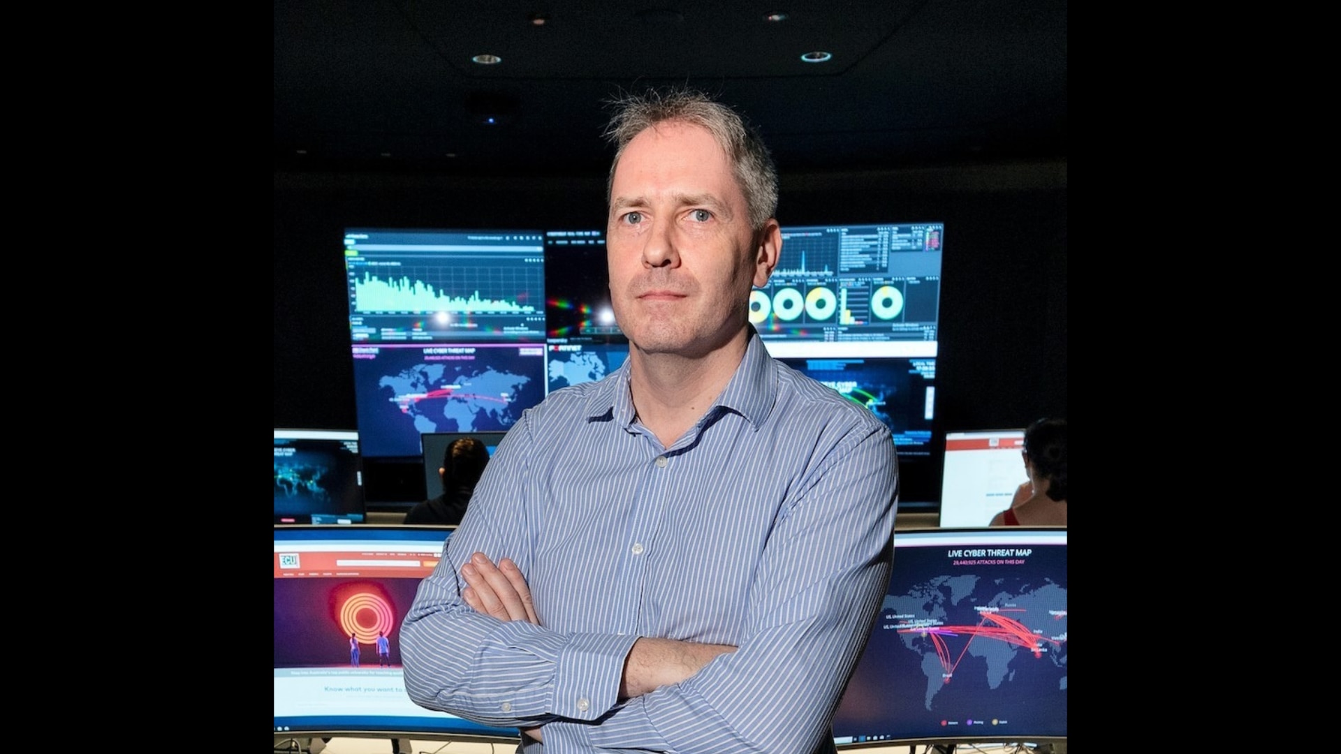 A middle-aged man with grey hair stands with arms folded in front of a bank of computer monitors.