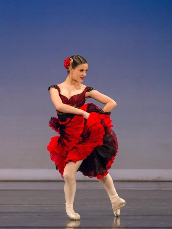A young girl in a red ballet dress and pointe shoes performs on stage.