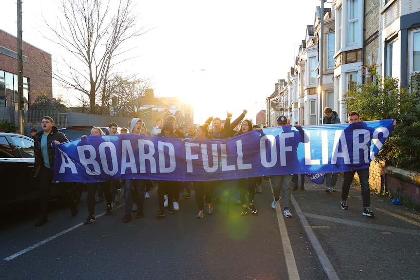 Everton fans walk down a street carrying a large banner that reads "A BOARD FULL OF LIARS"