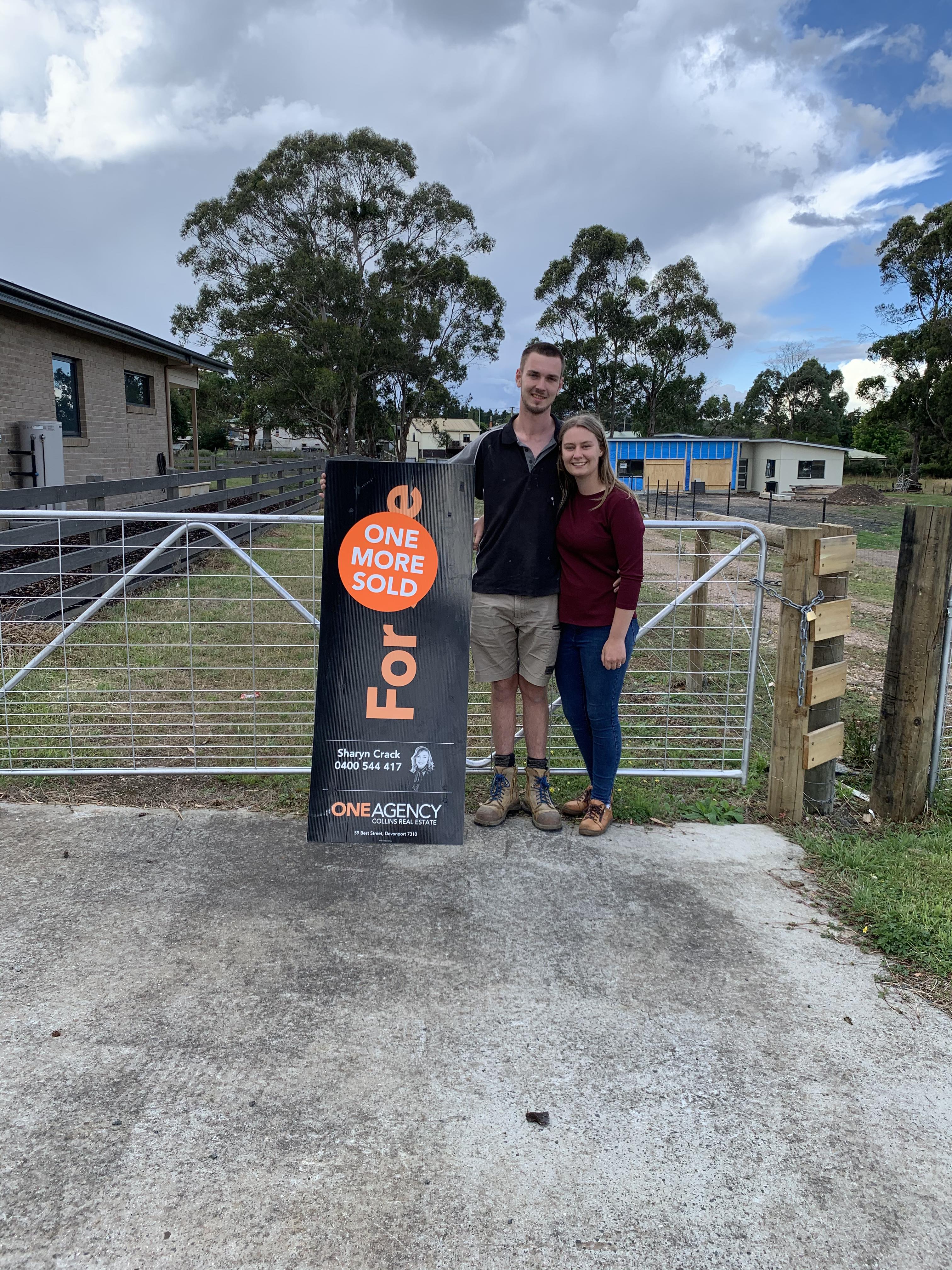 A young couple stand in front of a gate, with a "sold" real estate sign in front of them