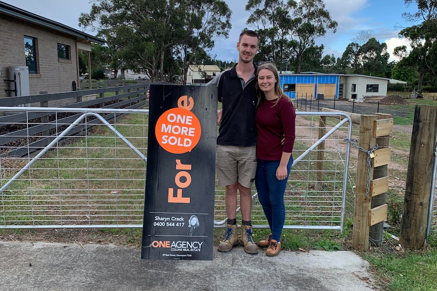 A young couple stand in front of a gate, with a "sold" real estate sign in front of them