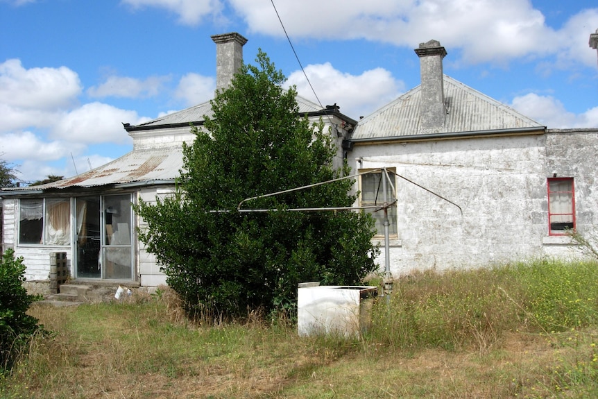 A broken down old house surrounded by long grass. Broken hills hoist in foreground