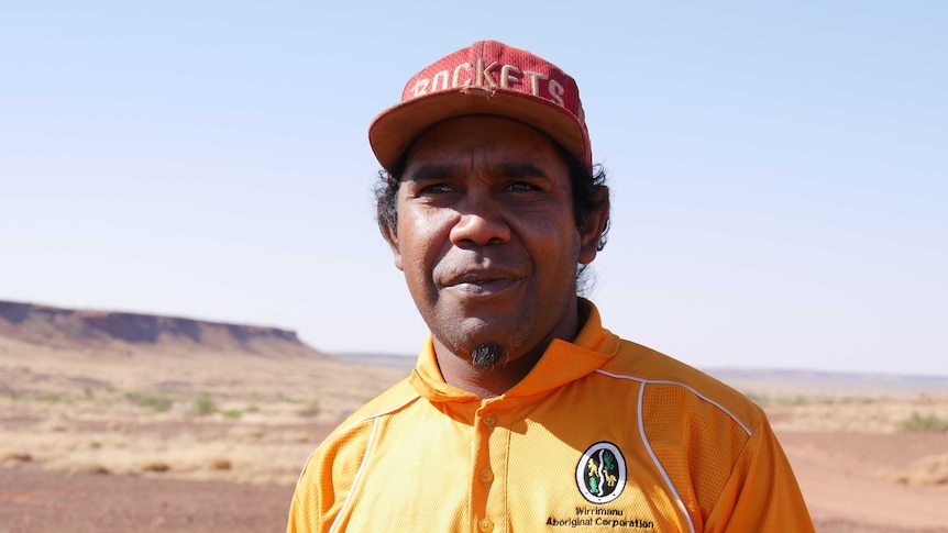 An Indigenous man pictured outside in the desert. He wears a red cap and a yellow Wirrimanu Aboriginal Corporation t-shirt.