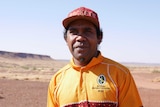An Indigenous man pictured outside in the desert. He wears a red cap and a yellow Wirrimanu Aboriginal Corporation t-shirt.