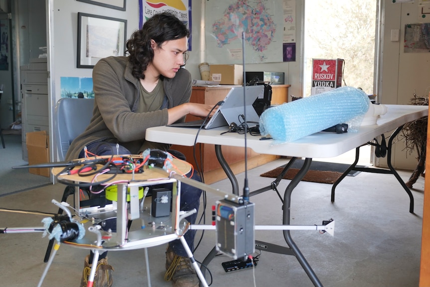 A young man with wavy short hair sits at a foldout table with his computer and a contraption next to him.