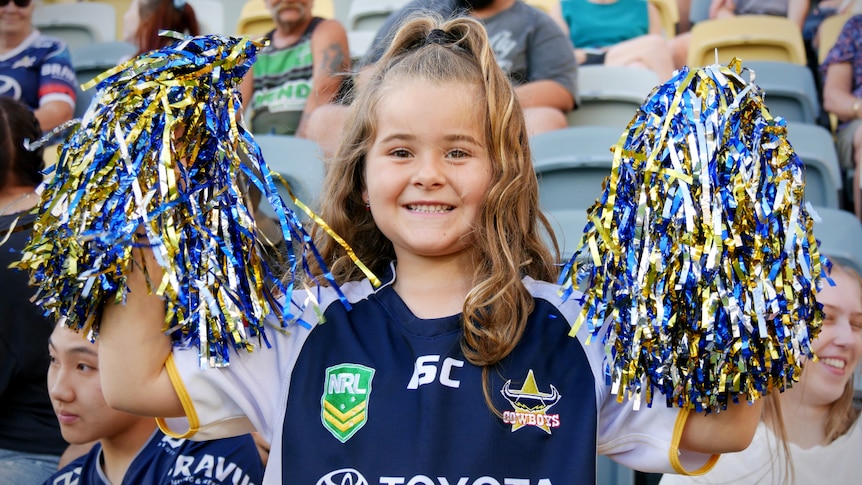 A young girl wearing a Cowboys jersey holds a pair of pompoms in a stadium grandstand 