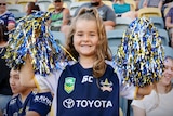 A young girl wearing a Cowboys jersey holds a pair of pompoms in a stadium grandstand 