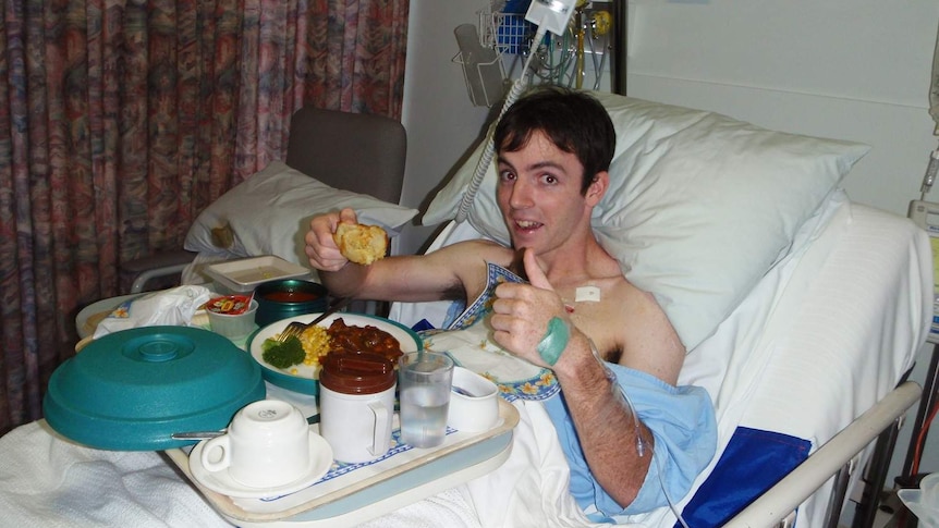 Dan Haslam lies in a hospital bed, smiling at the camera as he eats food from a tray.