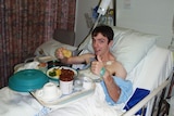 Dan Haslam lies in a hospital bed, smiling at the camera as he eats food from a tray.