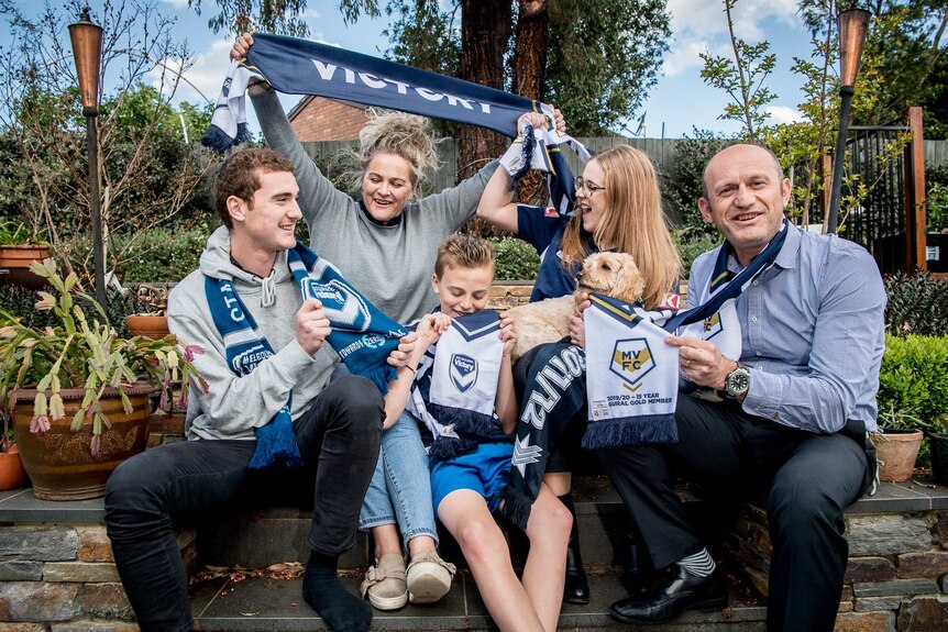 Five people sit on back steps. Mum holds up a Victory scarf.