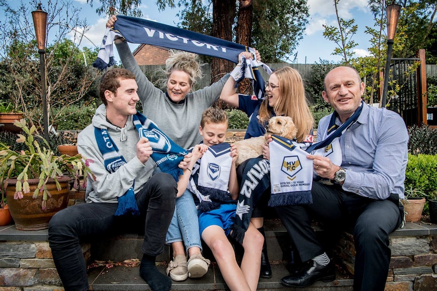 Five people sit on back steps. Mum holds up a Victory scarf.