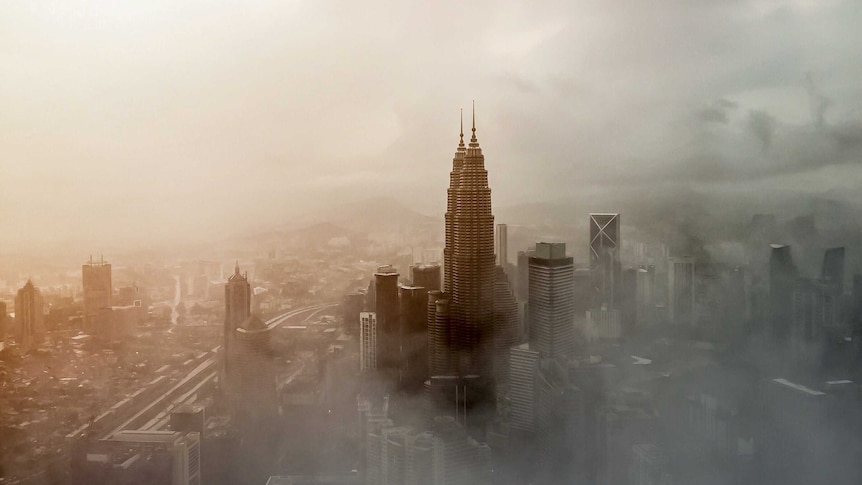 A high angle shows the twin Petronas Towers rising above fog and dominating the Kuala Lumpur skyline obscured by cloud.