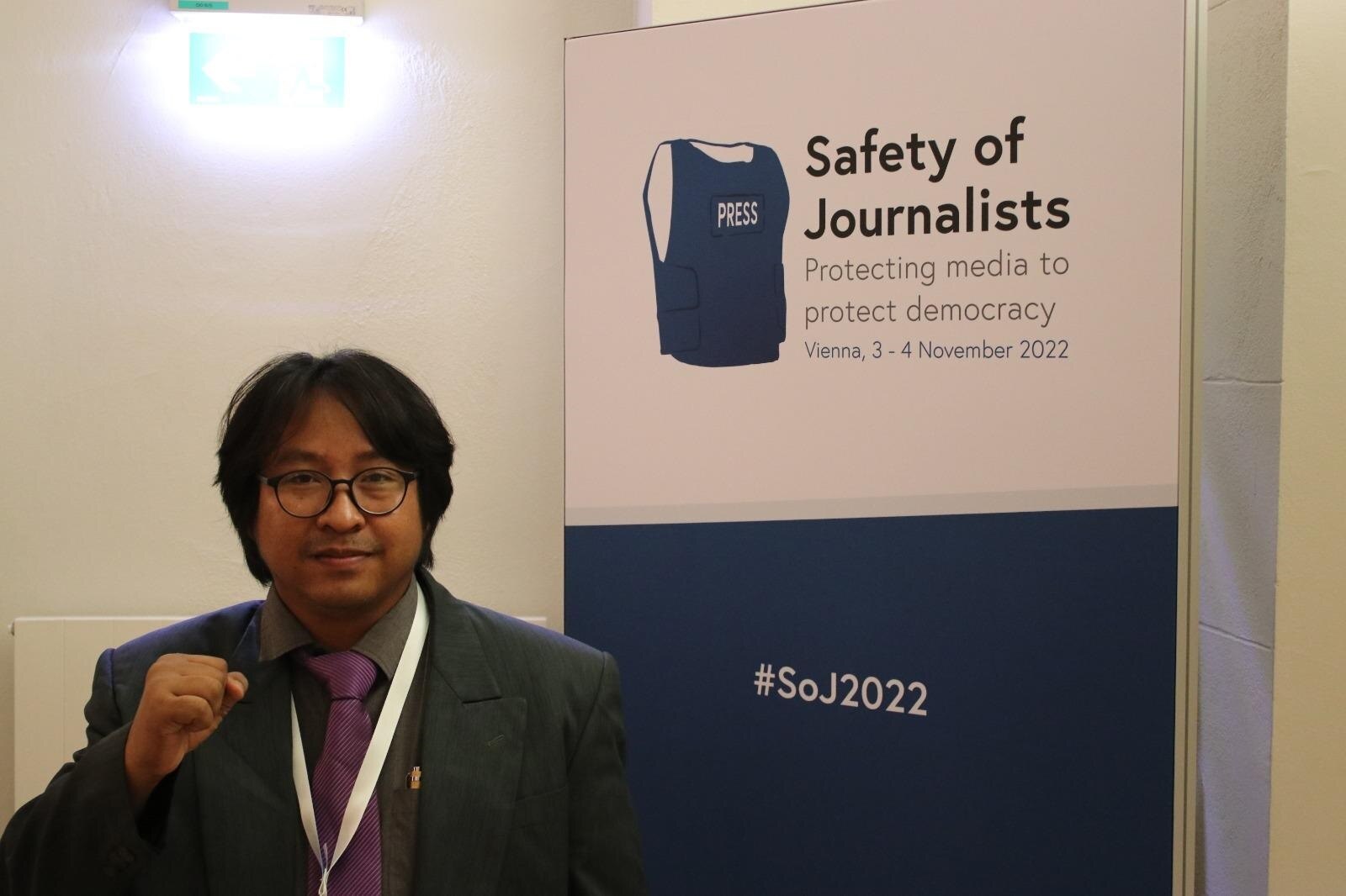 A suited man holds his right fist at chest height as he poses for a photo near a sign reading: "Safety of journalists."