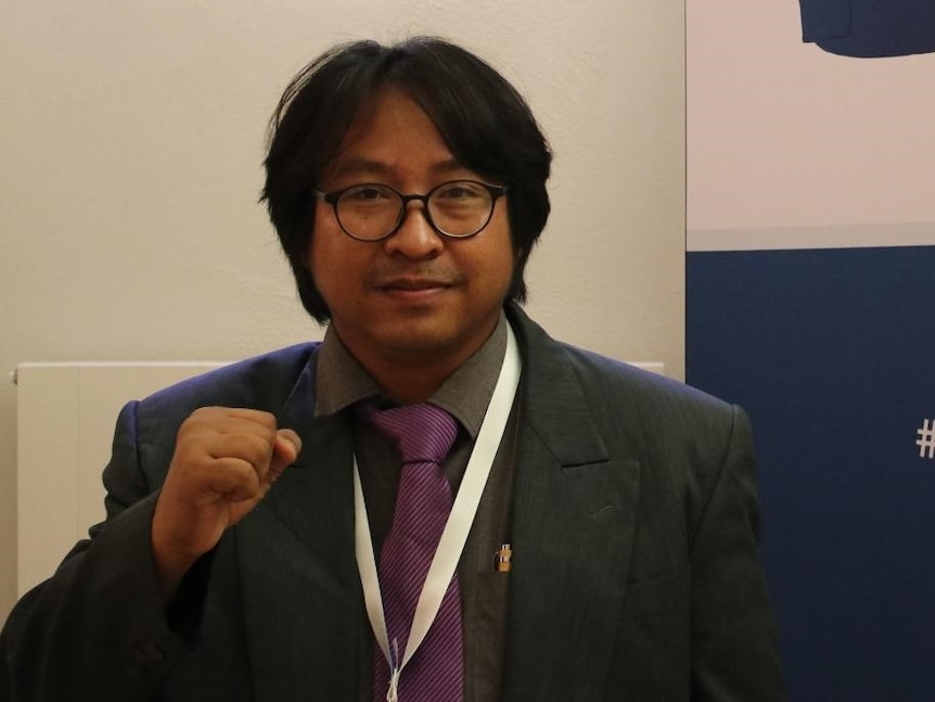 A suited man holds his right fist at chest height as he poses for a photo near a sign reading: "Safety of journalists."