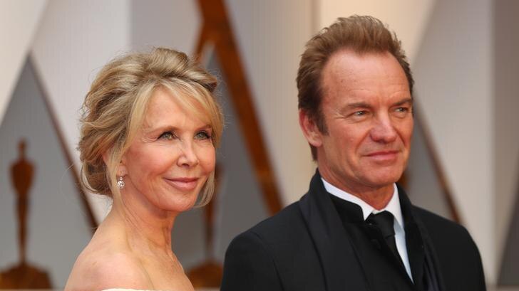 Trudie Styler, left, wears off the shoulder dress with her hair up as she stands next to Sting in black tuxedo. Both look away.