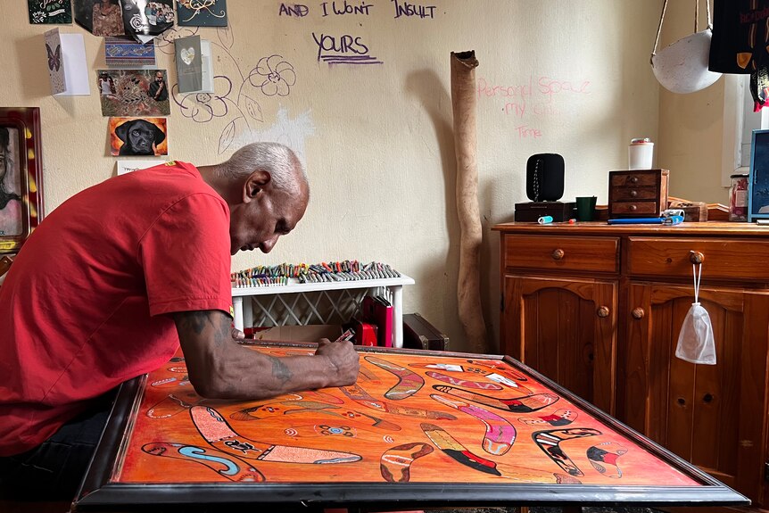 A man is bend over an Aboriginal artwork in a frame, adding the finishing touches.