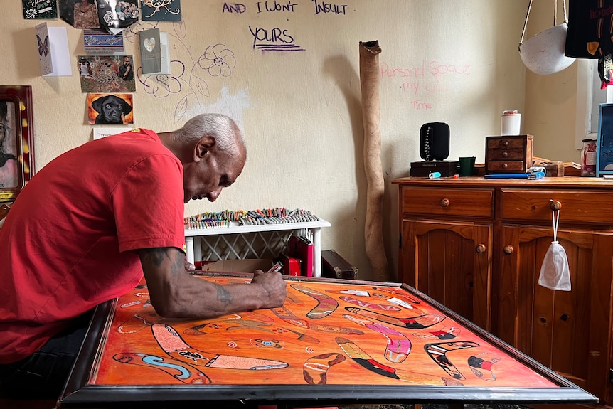 A man is bend over an Aboriginal artwork in a frame, adding the finishing touches.