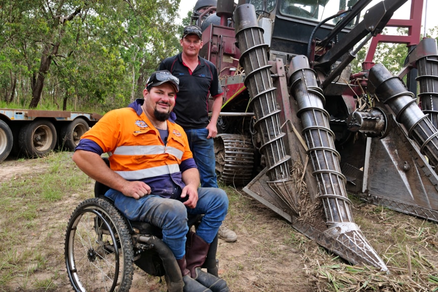 a man using a wheelchair is next to a man standing, both are smiling at the camera. a large harvester looms next to them