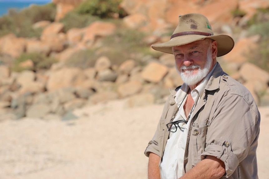 a man in a broad hat and white beard stands in a sandy outback area looking at the camera